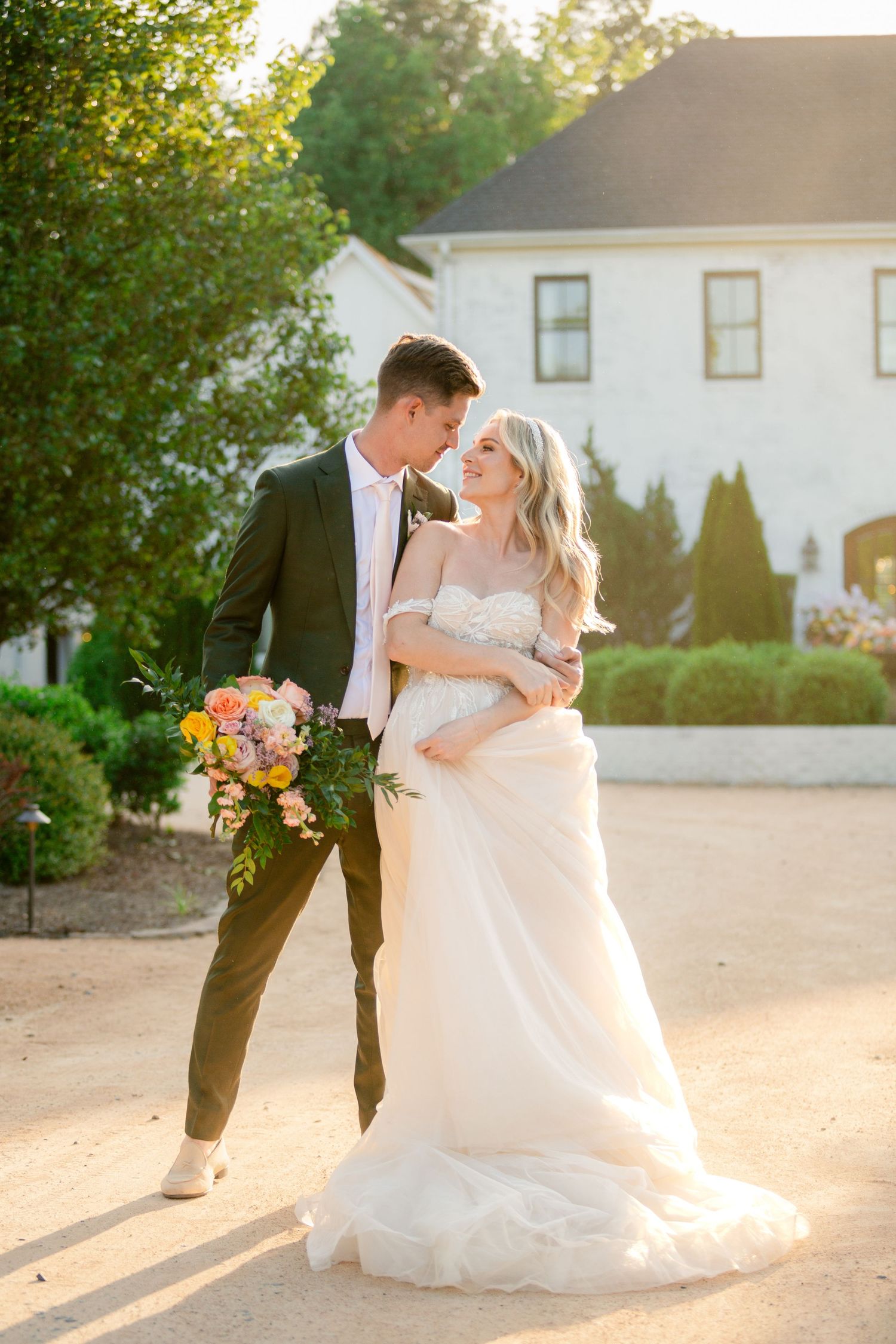 Newlywed couple embraces in romantic sunset moment outside white house with colorful summer bouquet.