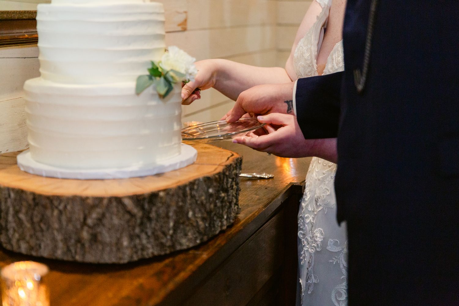Hands carefully cutting into an elegant white wedding cake displayed on a rustic wooden slice stand.