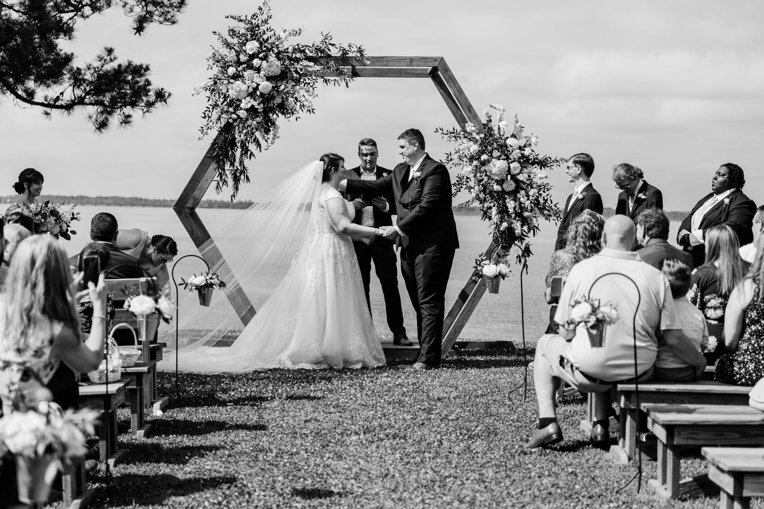 Black and white panoramic photo sequence showing a wedding ceremony taking place outdoors under a tree by the ocean.