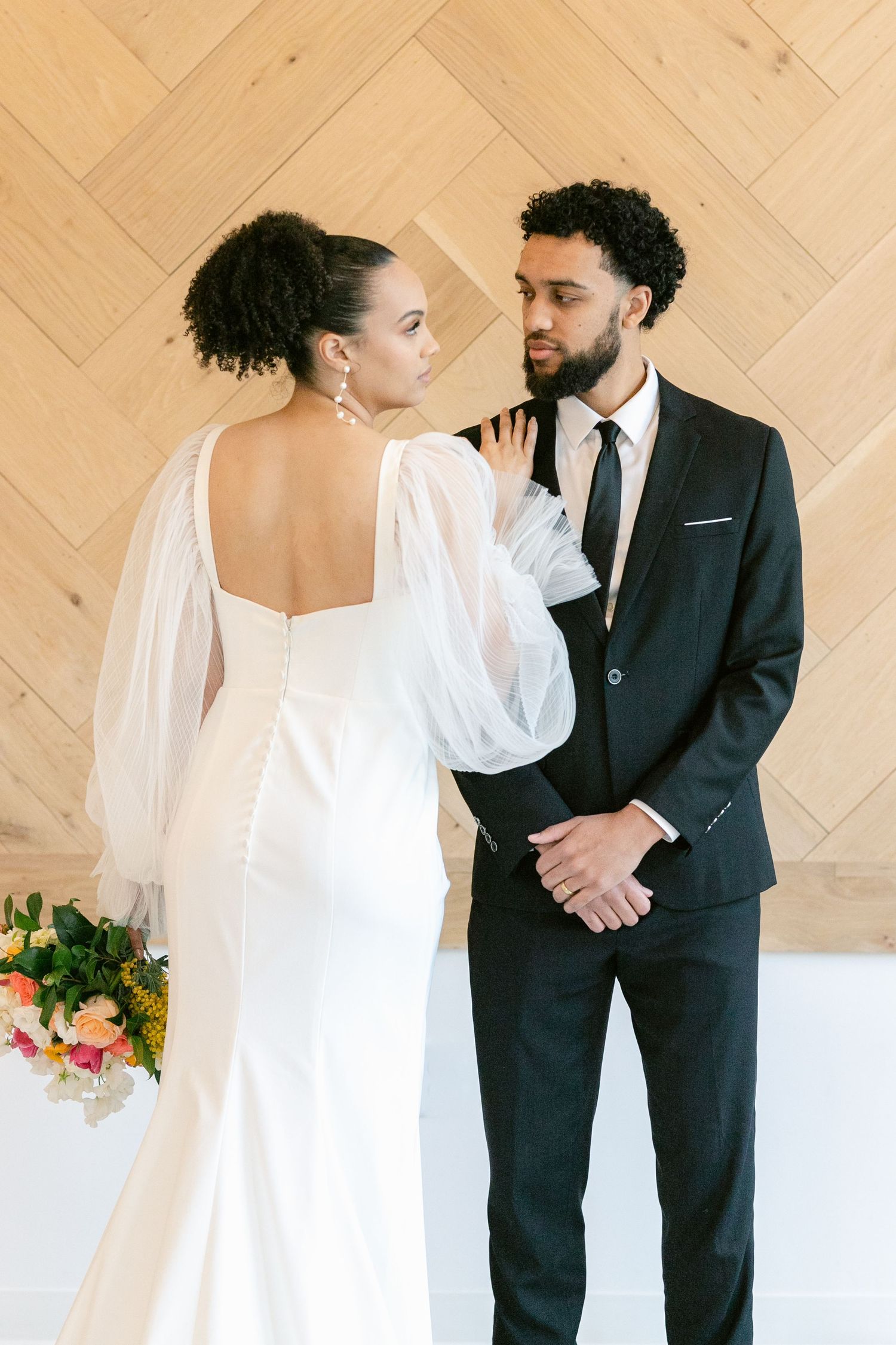 A romantic wedding photoshoot series showing a couple in white dress and black suit against a light wooden wall background.