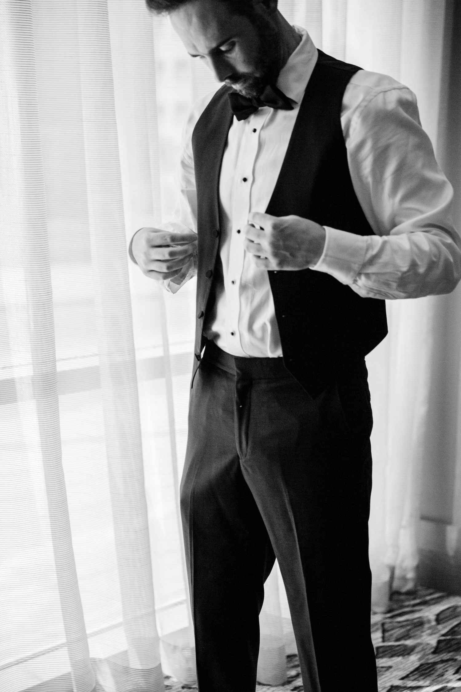 A person in formal attire adjusts their cufflinks while standing near white curtained windows.