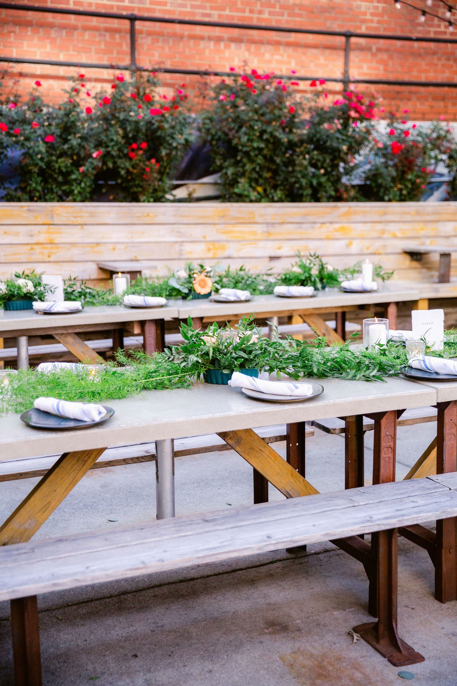 Long wooden picnic tables with benches arranged in a row on a patio with red brick walls and blooming flowers above.