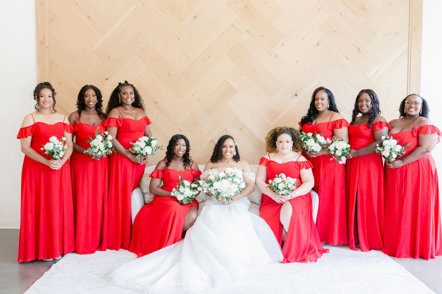 Bridesmaids wearing bright red dresses pose with bride in white gown for formal wedding party photo.