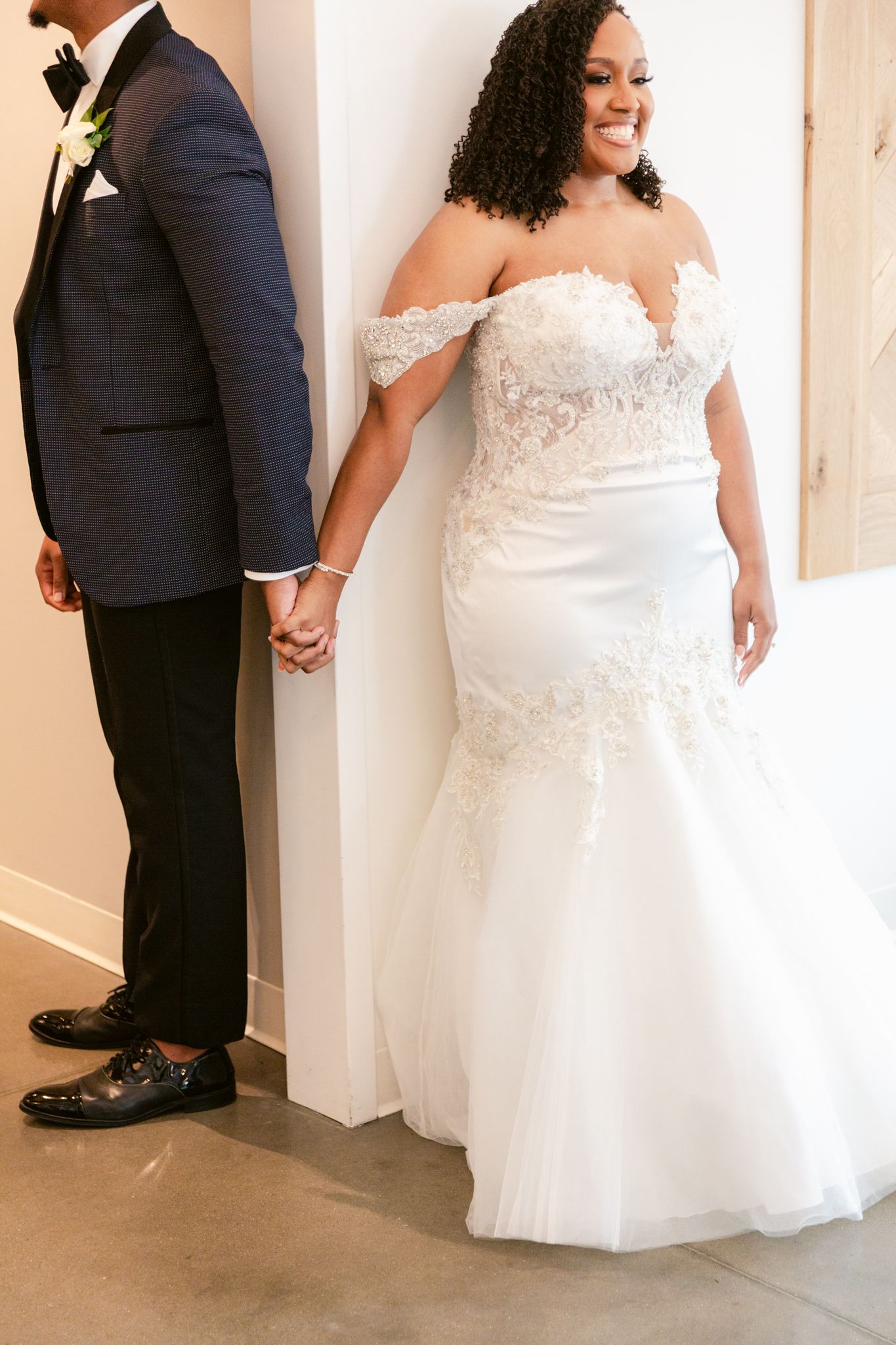 A bride in a white lace gown and groom in navy suit hold hands while standing against a light-colored wall.