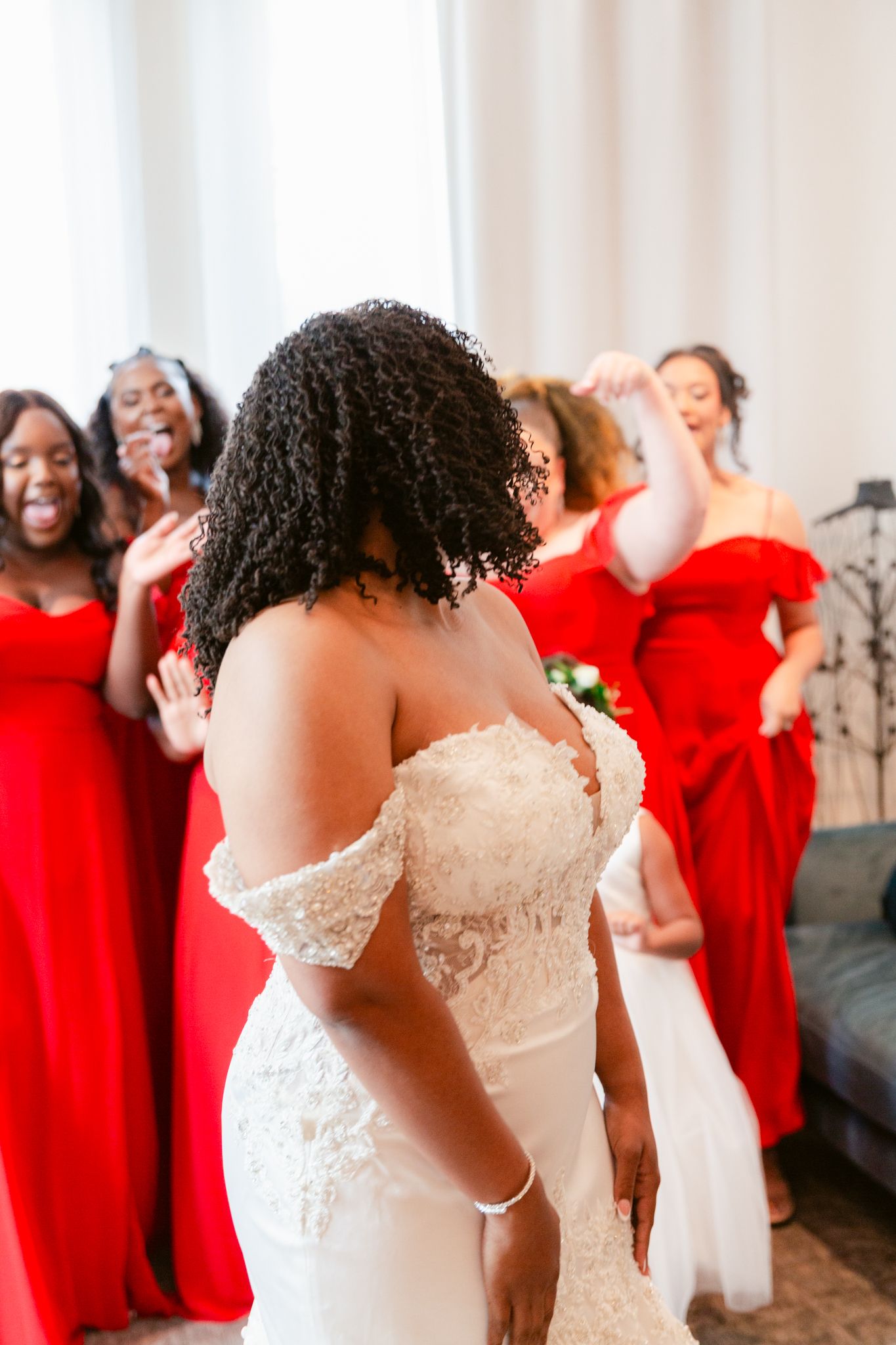 A bride in a white lace dress shares a joyful moment with bridesmaids wearing vibrant red gowns during wedding preparations.