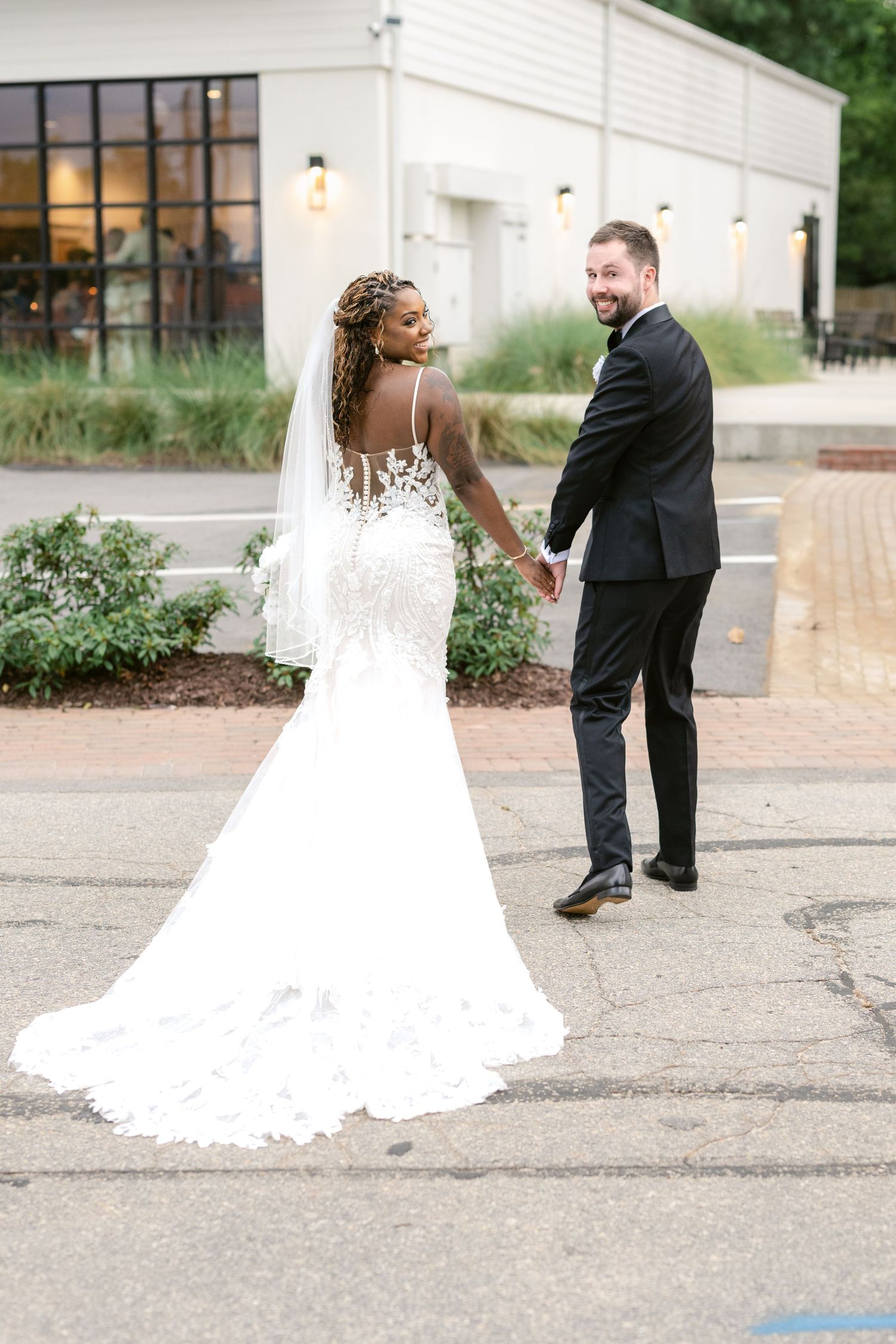 A newlywed couple shares a playful moment outside a modern white building with large windows.