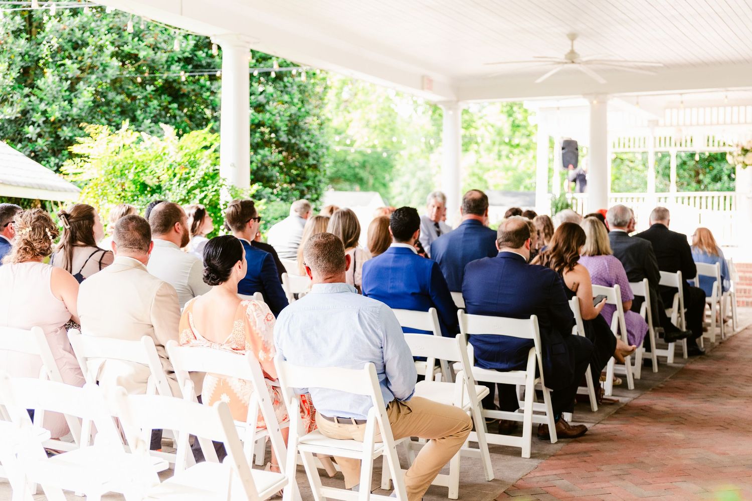 Wedding guests seated in white chairs under an outdoor covered pavilion during an outdoor ceremony.