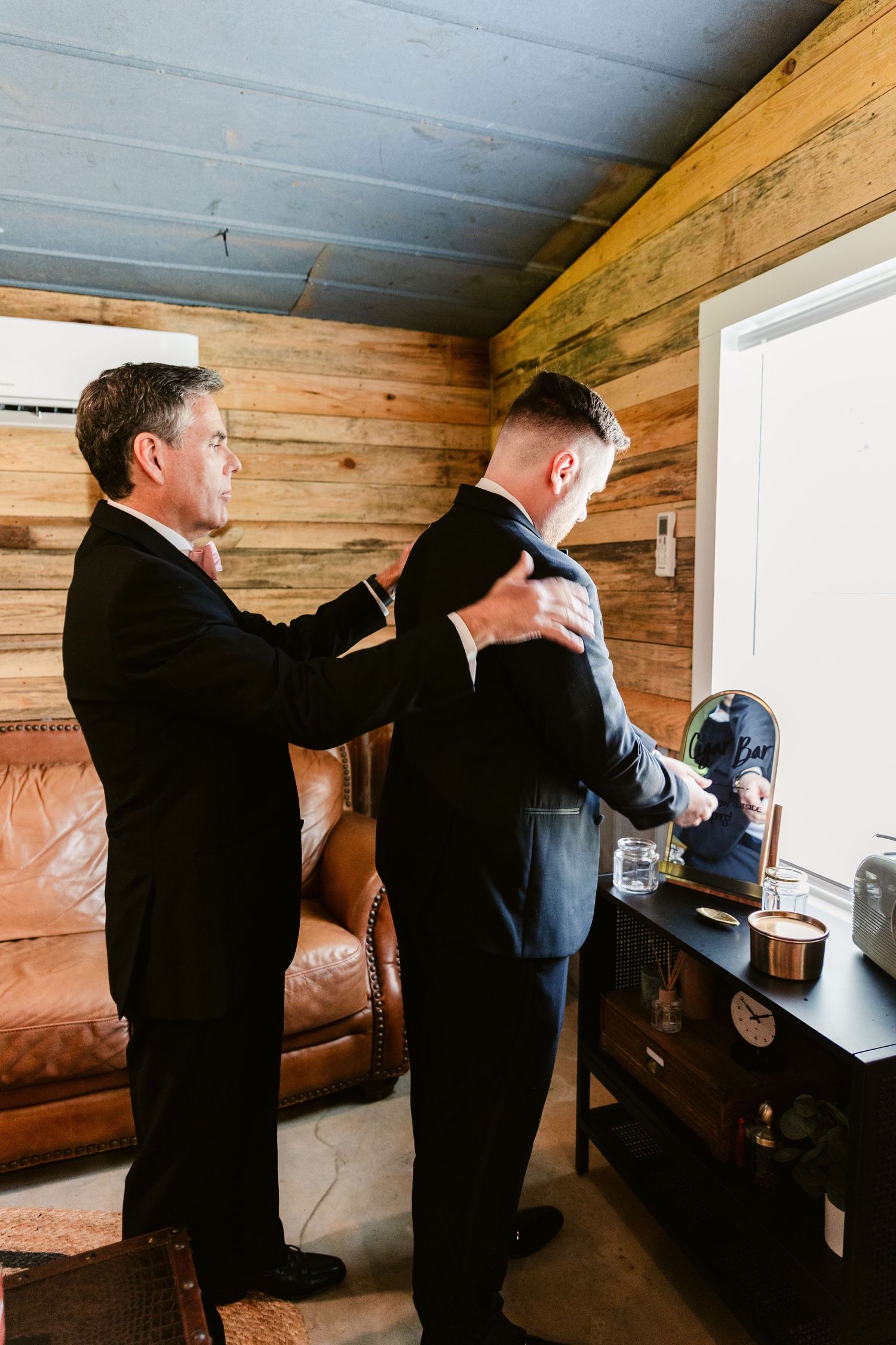 Two people in dark suits work together to install a window frame inside a rustic wooden room.