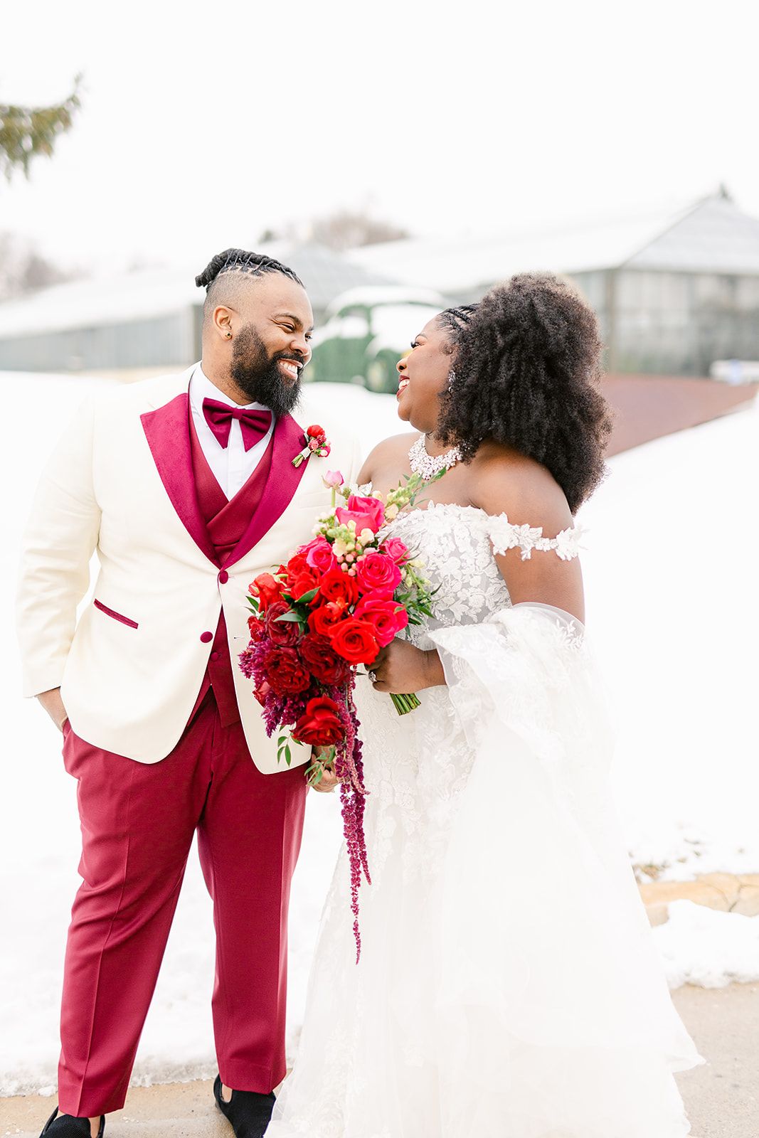 A joyful couple shares a candid moment on their wedding day, with the bride holding a vibrant red and pink bouquet.