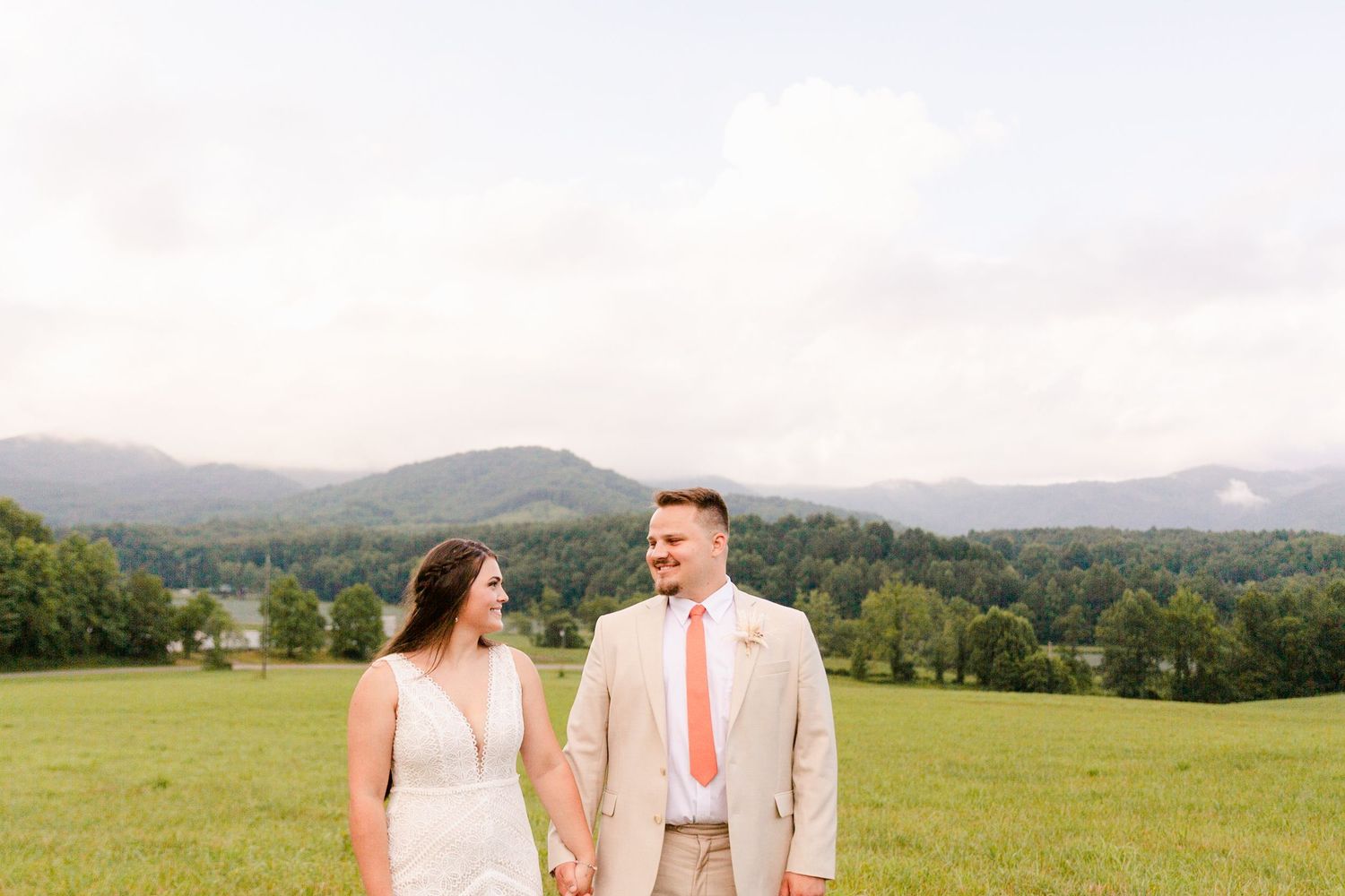 A couple shares a romantic moment in a scenic mountain meadow wearing light pink and beige attire.