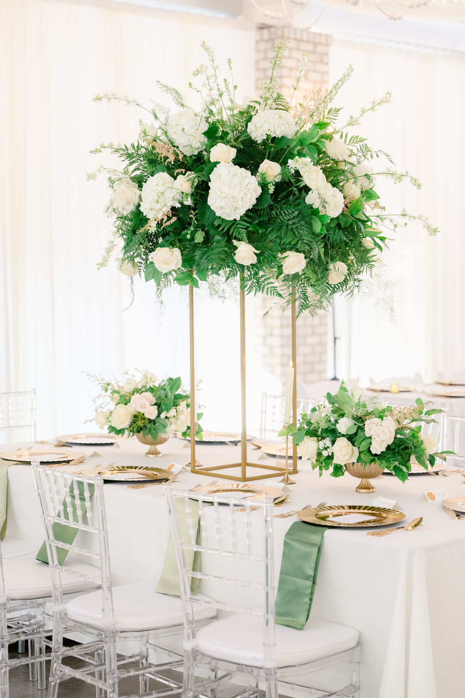 Elegant white and green wedding reception tables with tall floral centerpieces and gold accents in a bright venue.