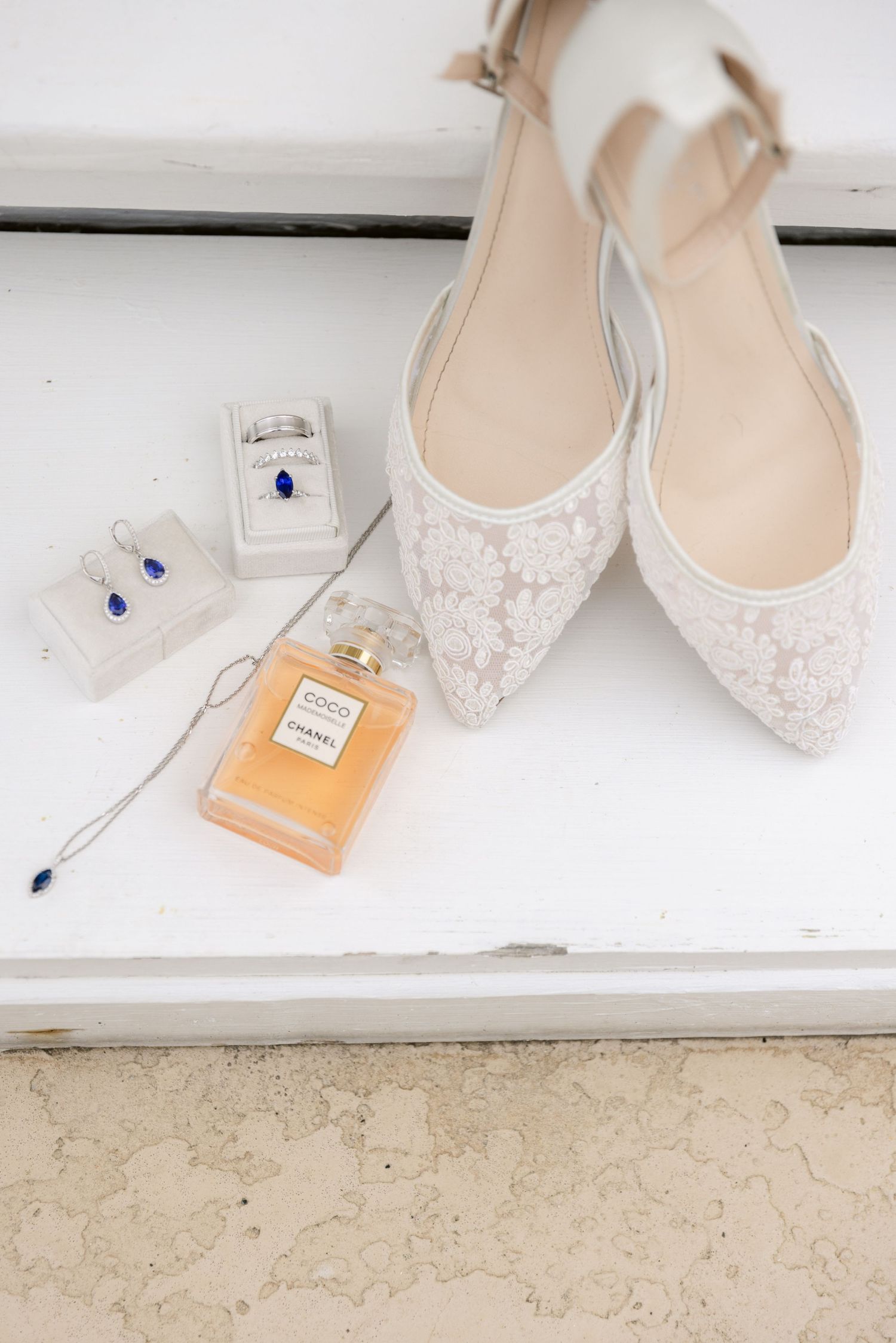Delicate white lace bridal flats displayed with perfume bottle and jewelry on white surface.