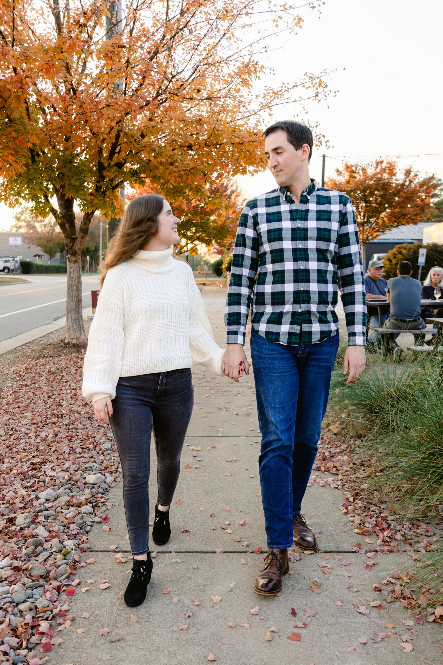 A couple takes a romantic autumn stroll on a sidewalk lined with orange trees and fallen leaves.