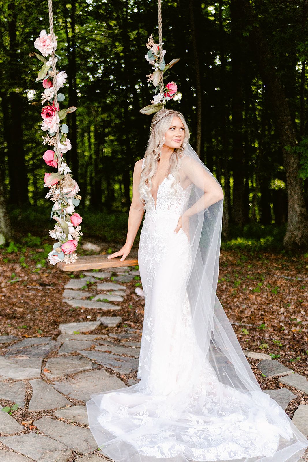 Bride in lace gown poses near floral swing in enchanted forest setting.