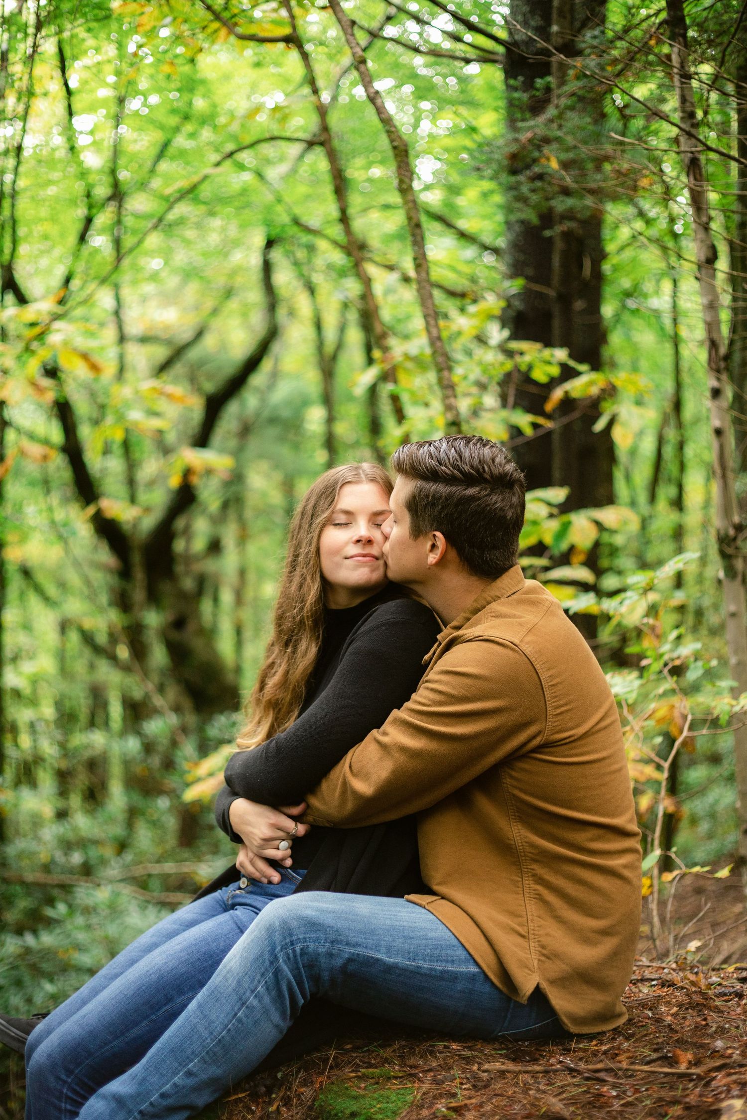 Intimate embrace between couple sitting on forest ground surrounded by green foliage.