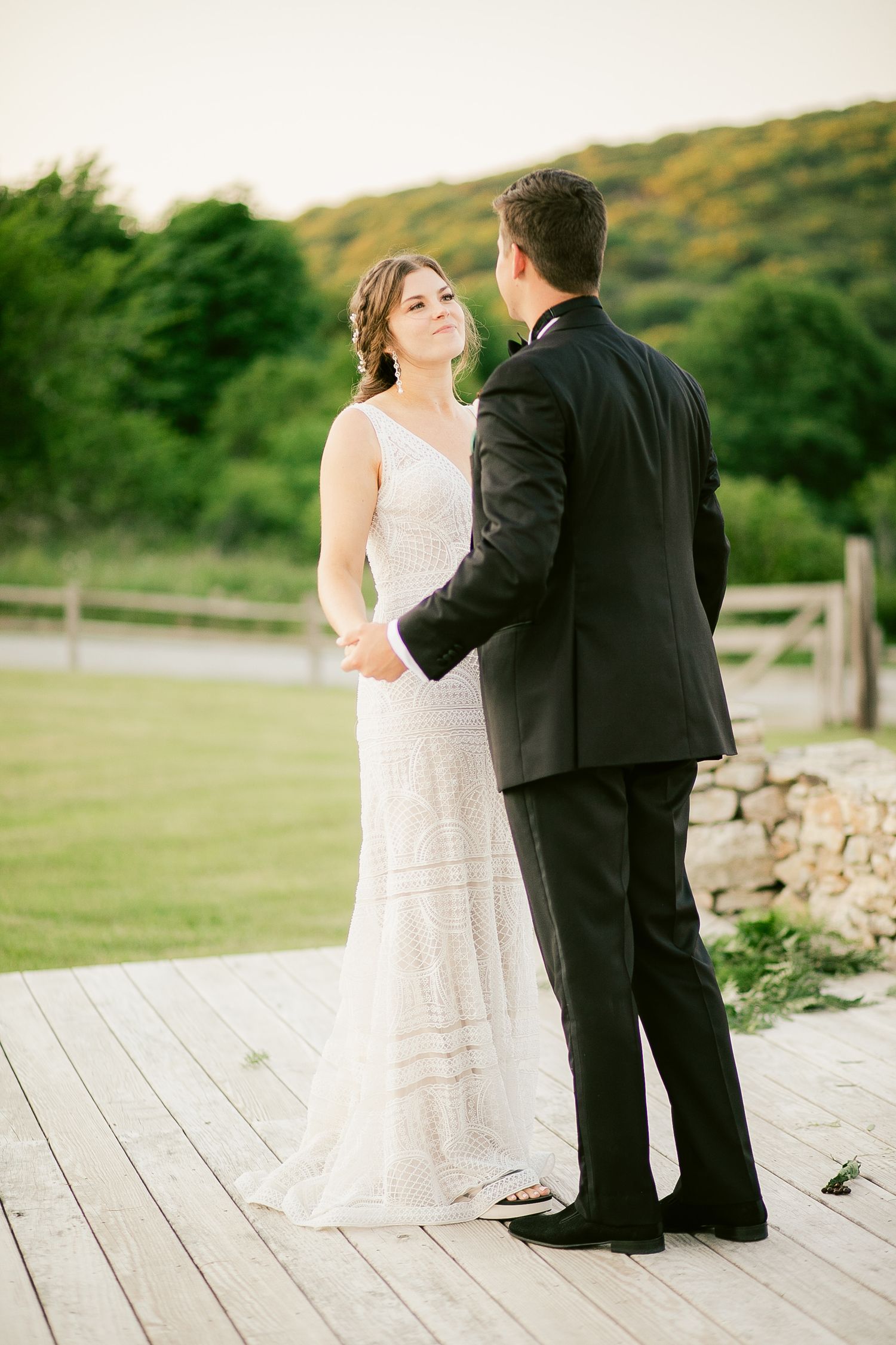 Bride in lace gown and groom in tuxedo share a moment on a wooden deck with mountain views.