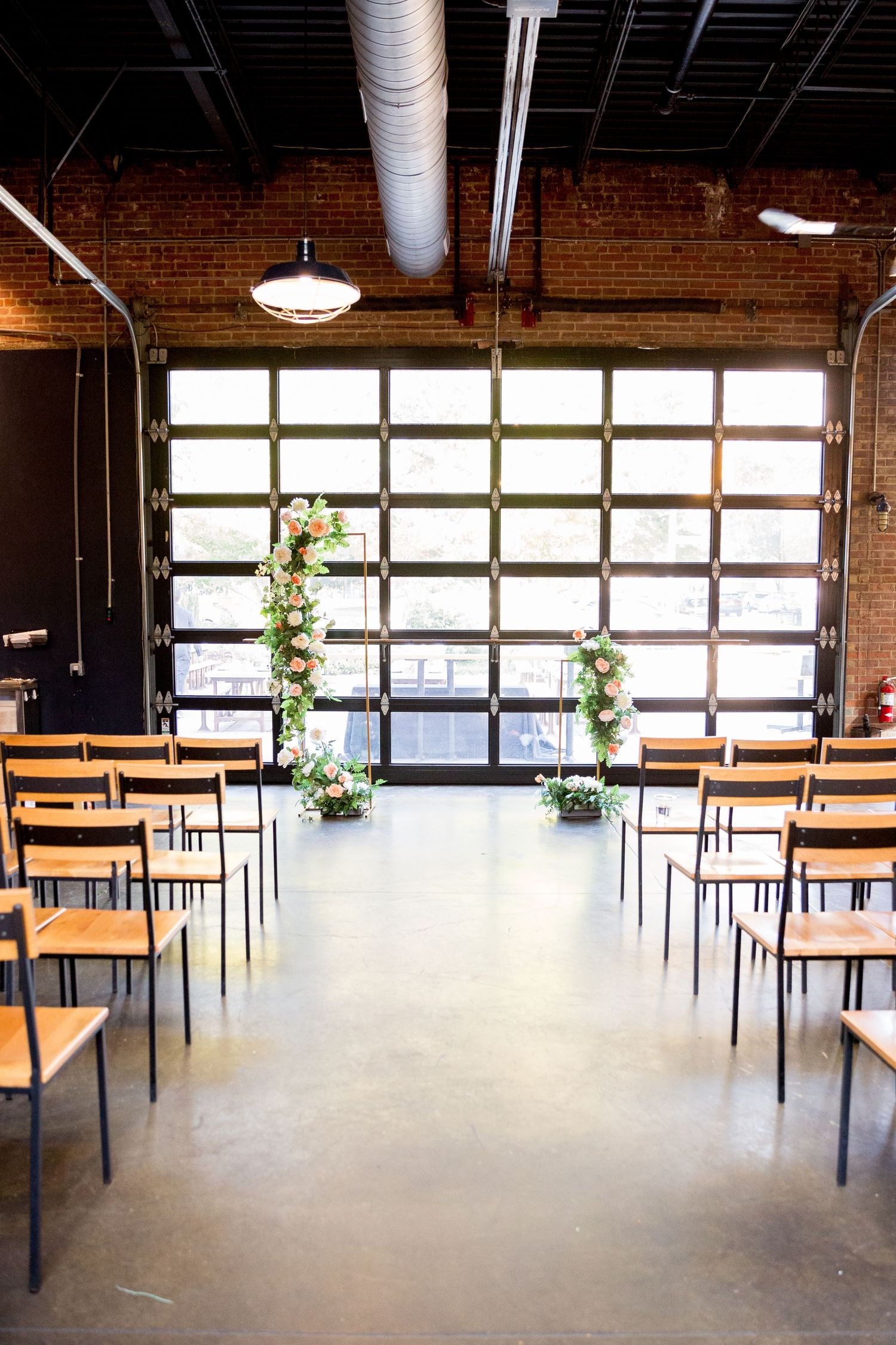 Industrial event space with wooden benches, exposed brick walls, and hanging plants by large windows provides modern ambiance.