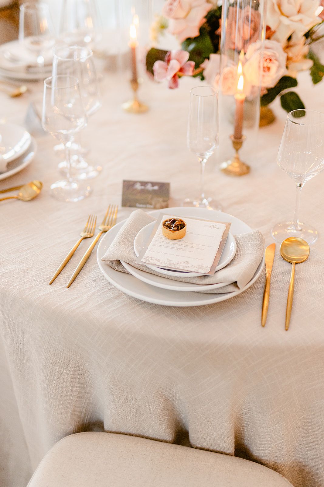 Formal place settings with gold flatware, white plates and blush roses on cream tablecloths.