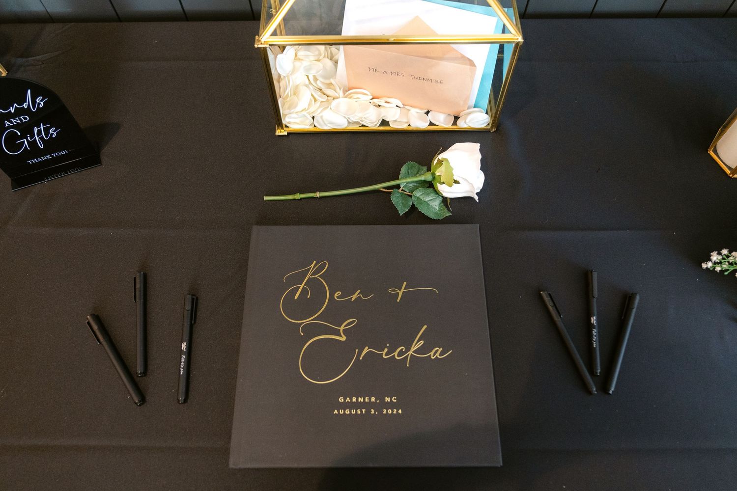 Black and gold wedding guest book with calligraphy names and pens on dark table with rose accent.
