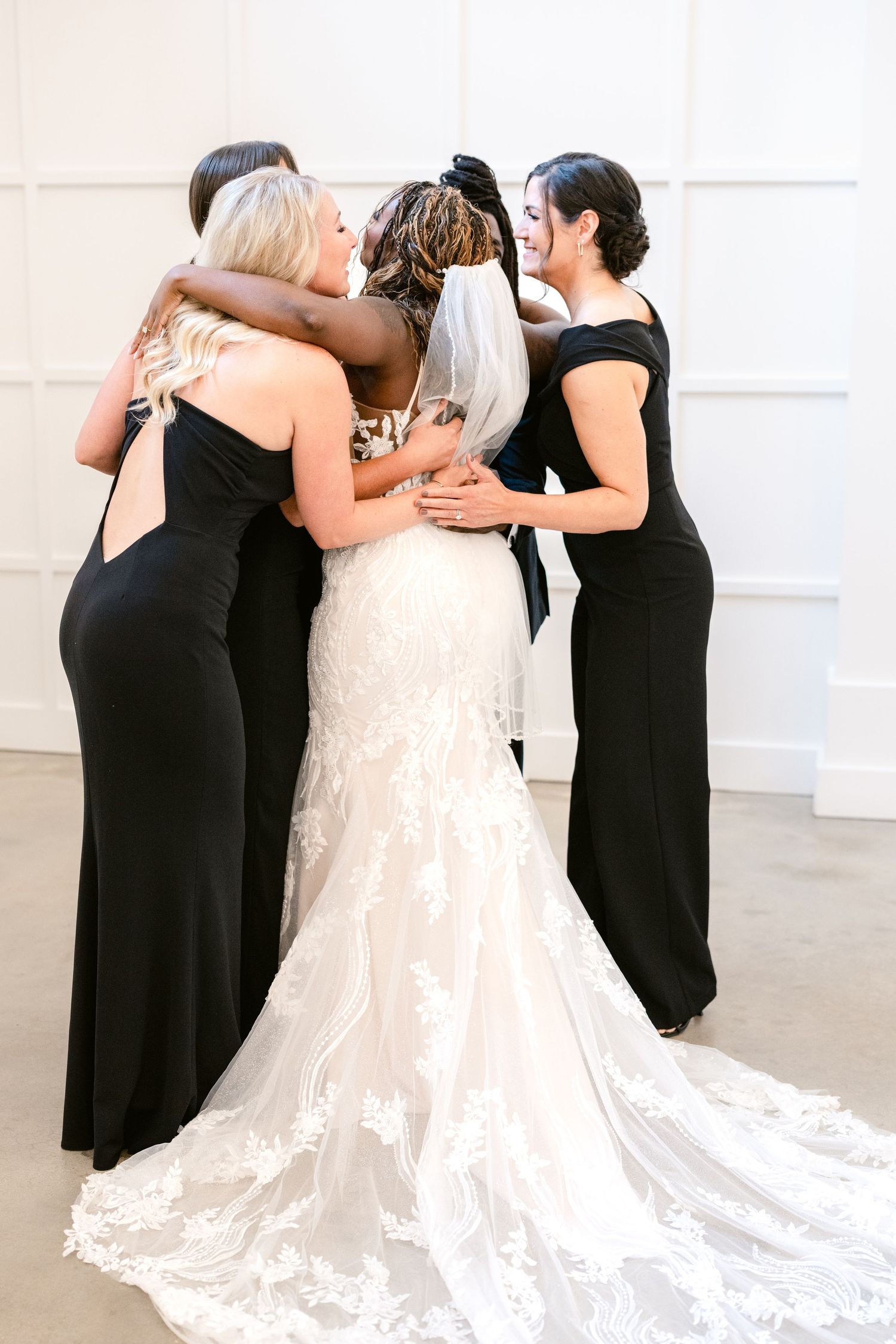 Bridesmaids in elegant black dresses embrace a bride wearing a stunning lace gown with a flowing train.