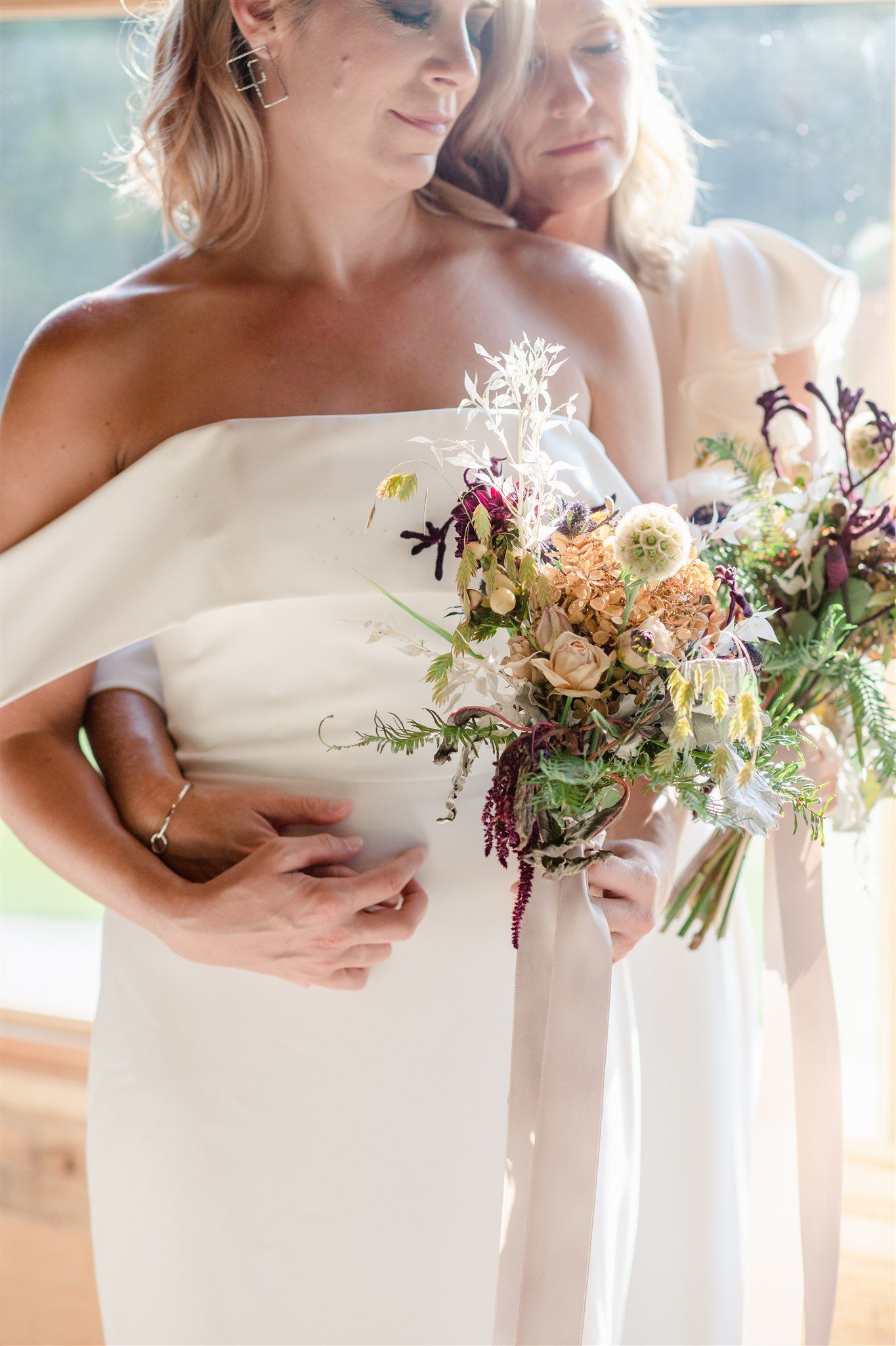 A wedding party in white dresses holds elegant wildflower bouquets with peach and purple blooms in natural light.
