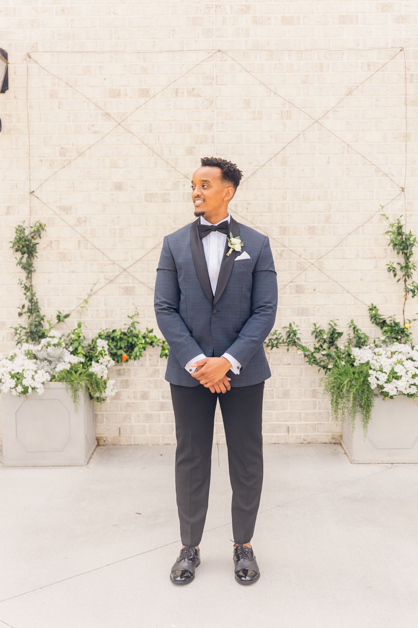 Groom in a charcoal suit and bowtie stands against white wall with climbing vines during wedding photoshoot.