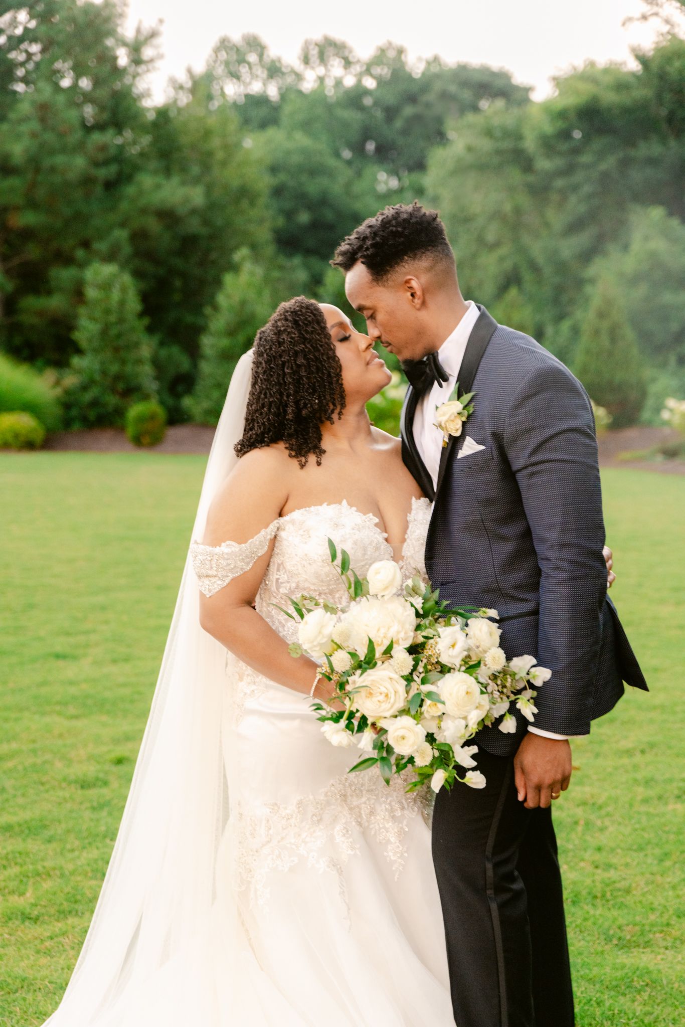 Newlywed couple shares romantic moments in a lush green outdoor setting with elegant white floral bouquet.