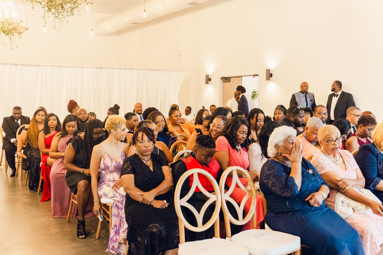 Wedding guests seated in rows watch attentively during an indoor ceremony at an elegant venue.