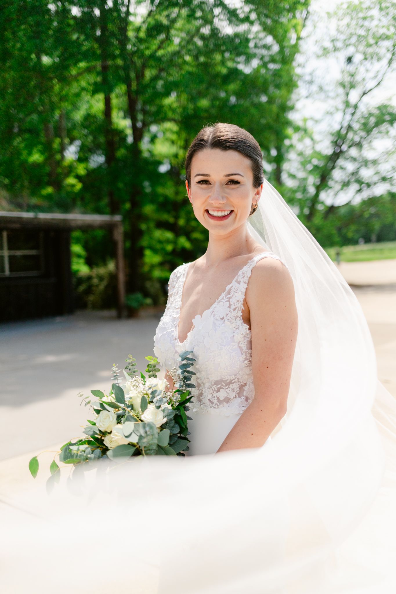A bride in a white lace wedding dress poses outdoors with a delicate bouquet of white and green flowers.