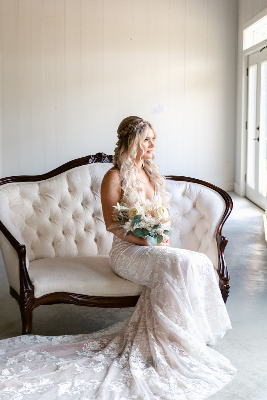 A bride in a lace wedding gown poses elegantly on a white tufted vintage settee in a bright sunlit room.