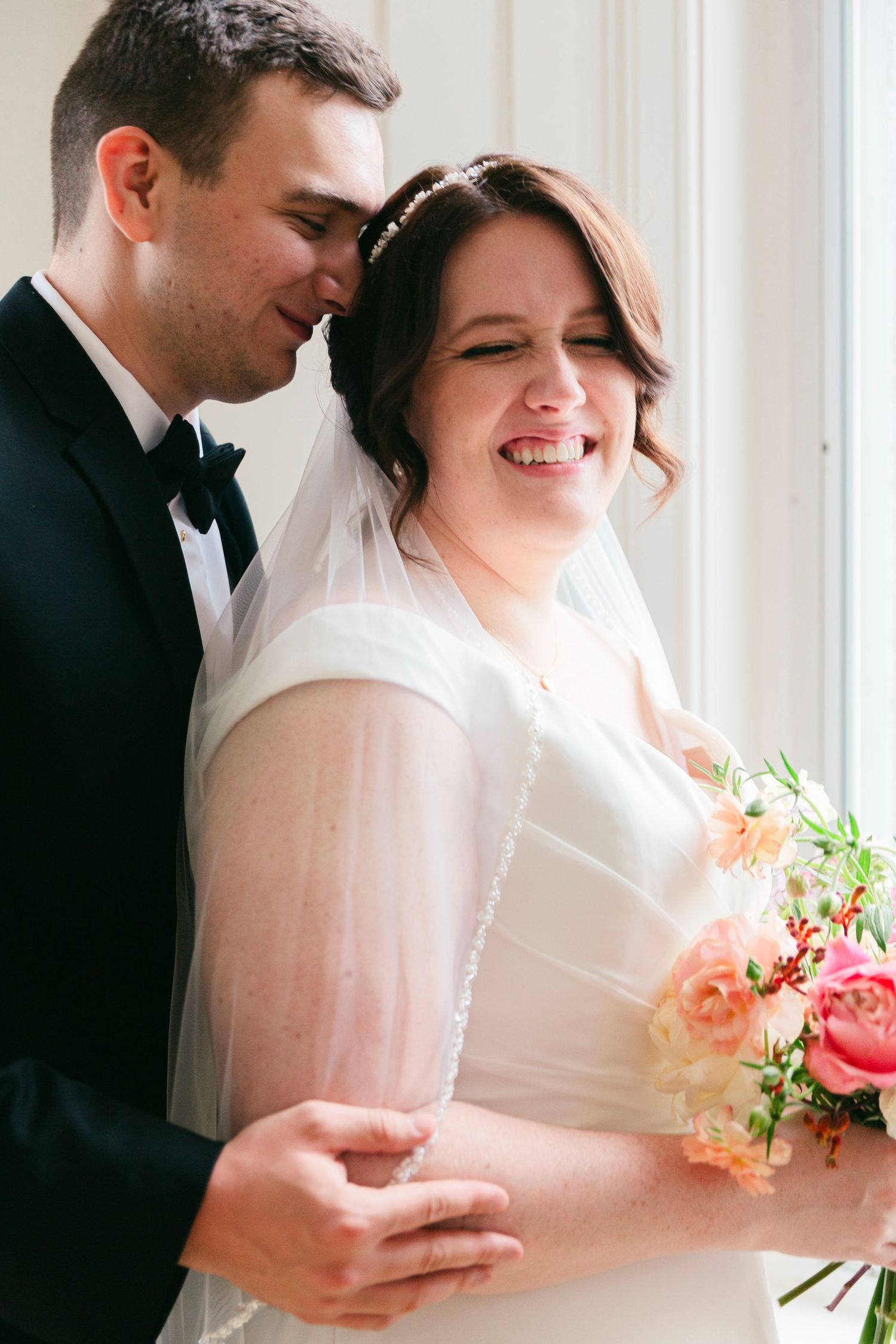 A romantic wedding moment captured in soft light as newlyweds share an intimate embrace while holding a coral bouquet.