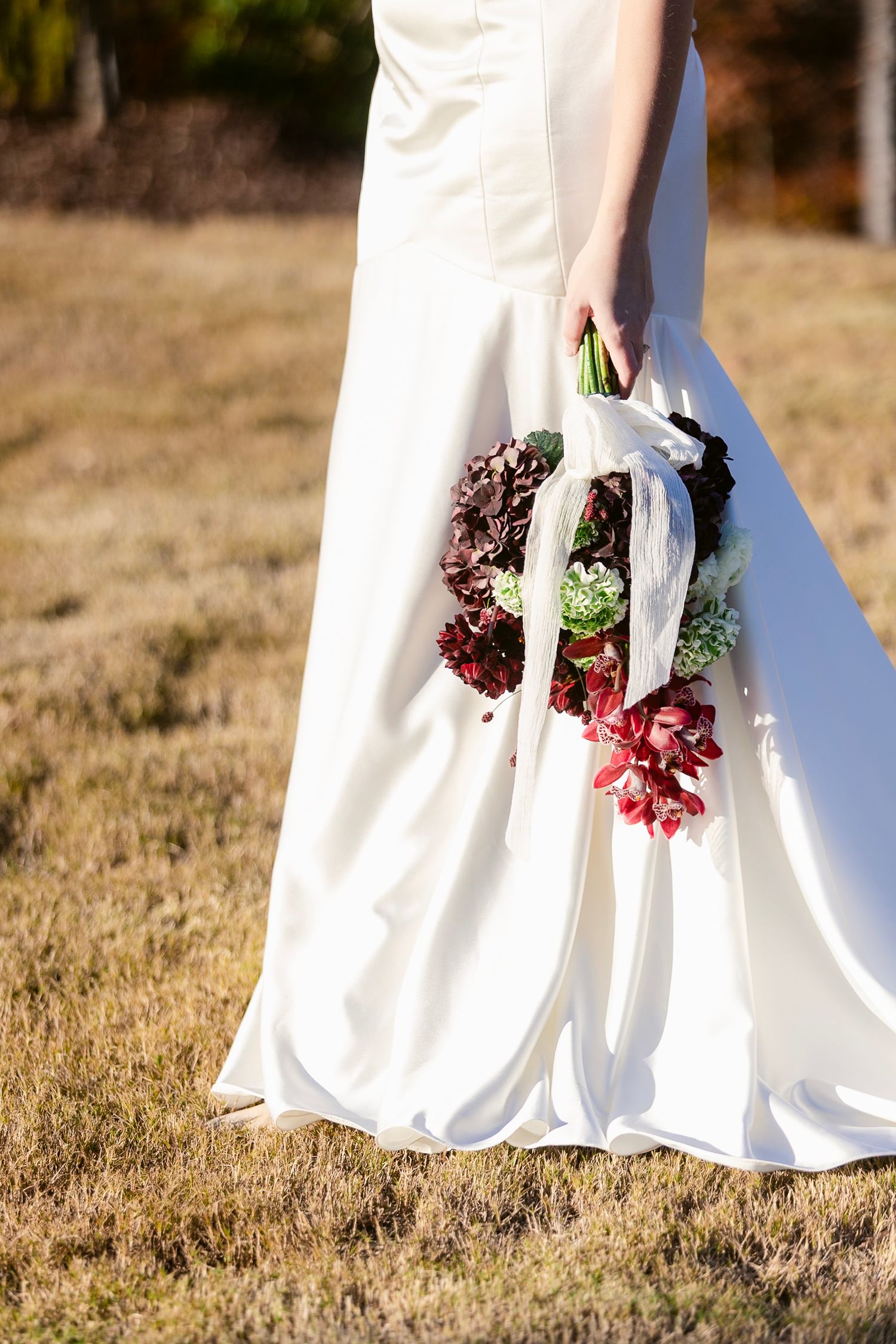 A bride in a flowing white wedding dress holds a vibrant bouquet of burgundy and pink flowers against a grassy field.