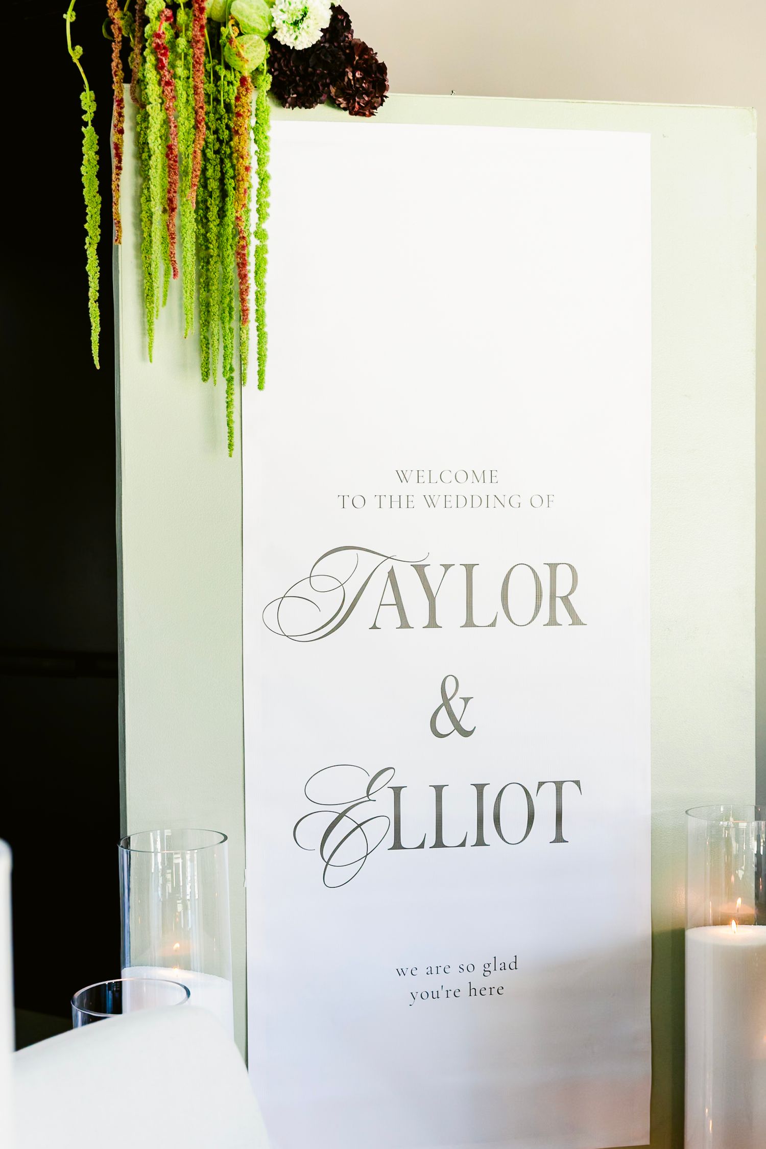 An elegant wedding welcome sign for Taylor & Elliot decorated with hanging greenery and surrounded by glass candle holders.
