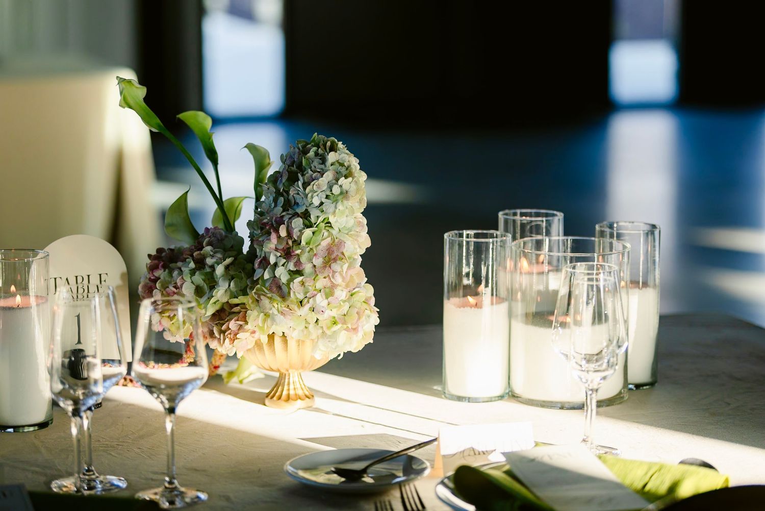 Elegant table settings with white hydrangeas and glassware in natural sunlight at an event venue.