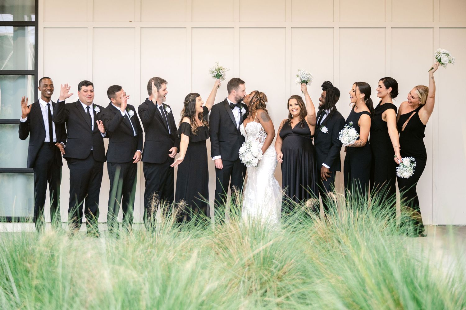 Wedding party stands in line against a modern building with tall grass in the foreground.