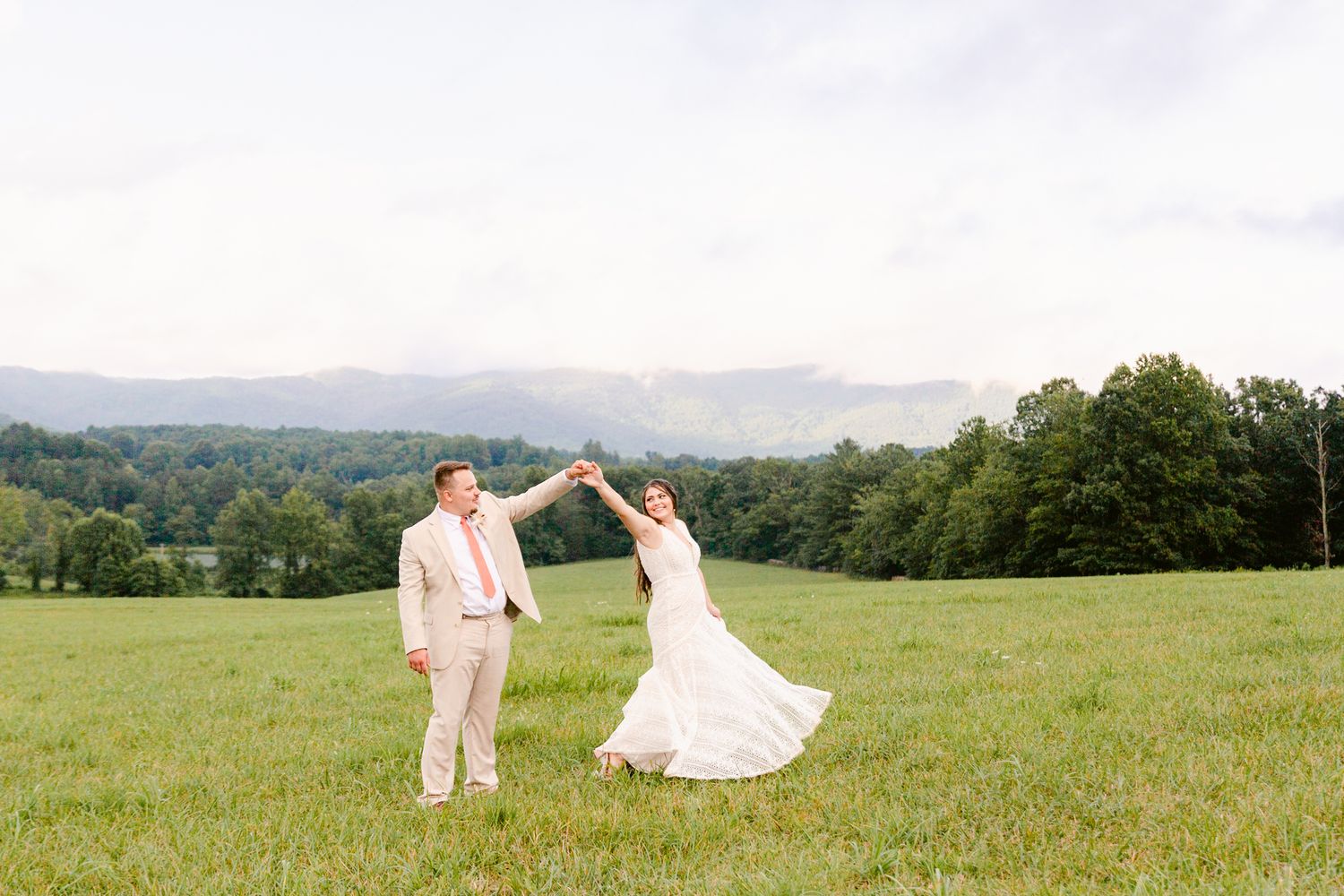 A couple in light-colored attire strolls hand in hand through a sunlit meadow with rolling hills beyond.