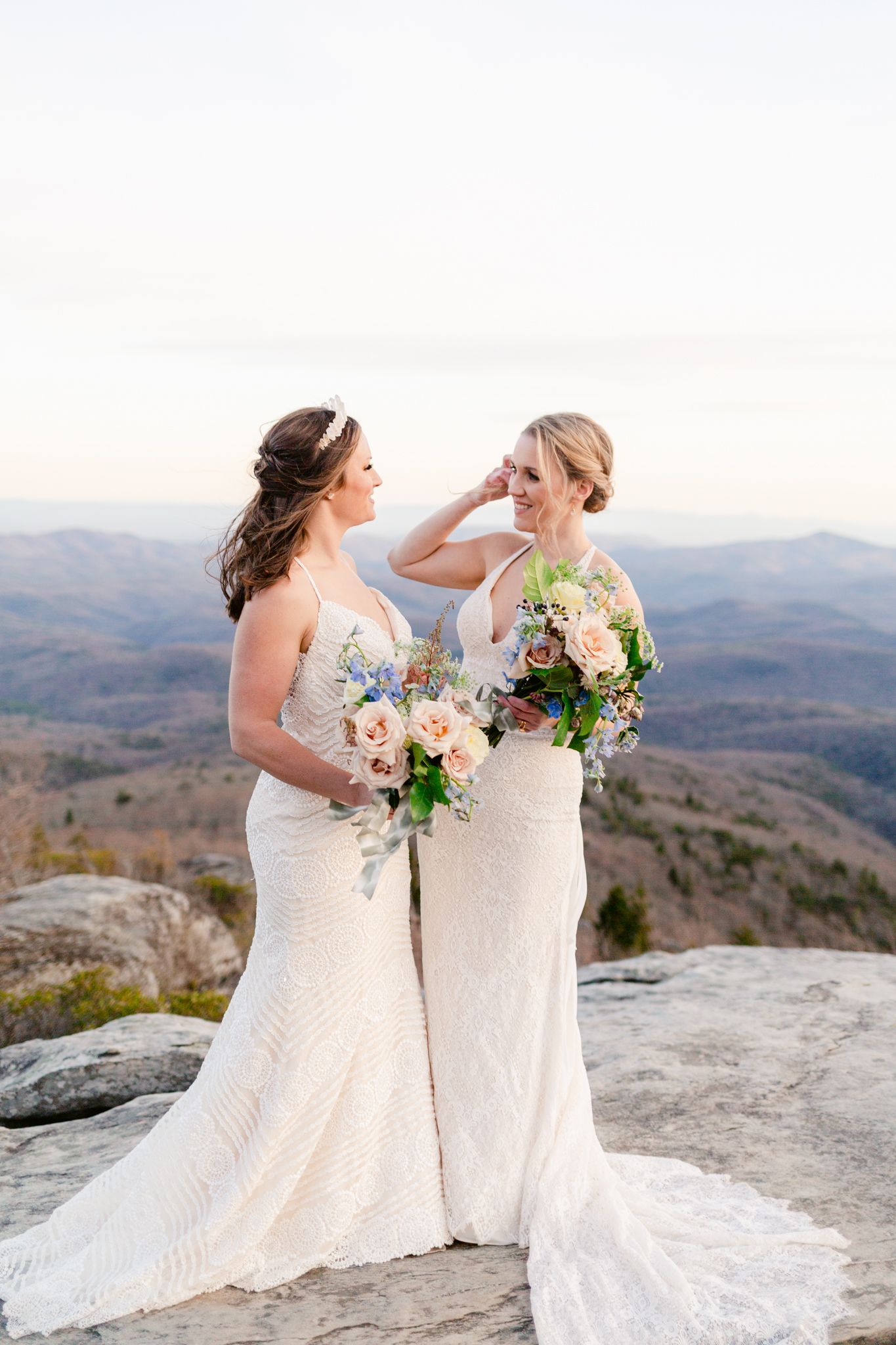 Two people in white wedding dresses share an intimate moment on a rocky mountain peak at dusk.