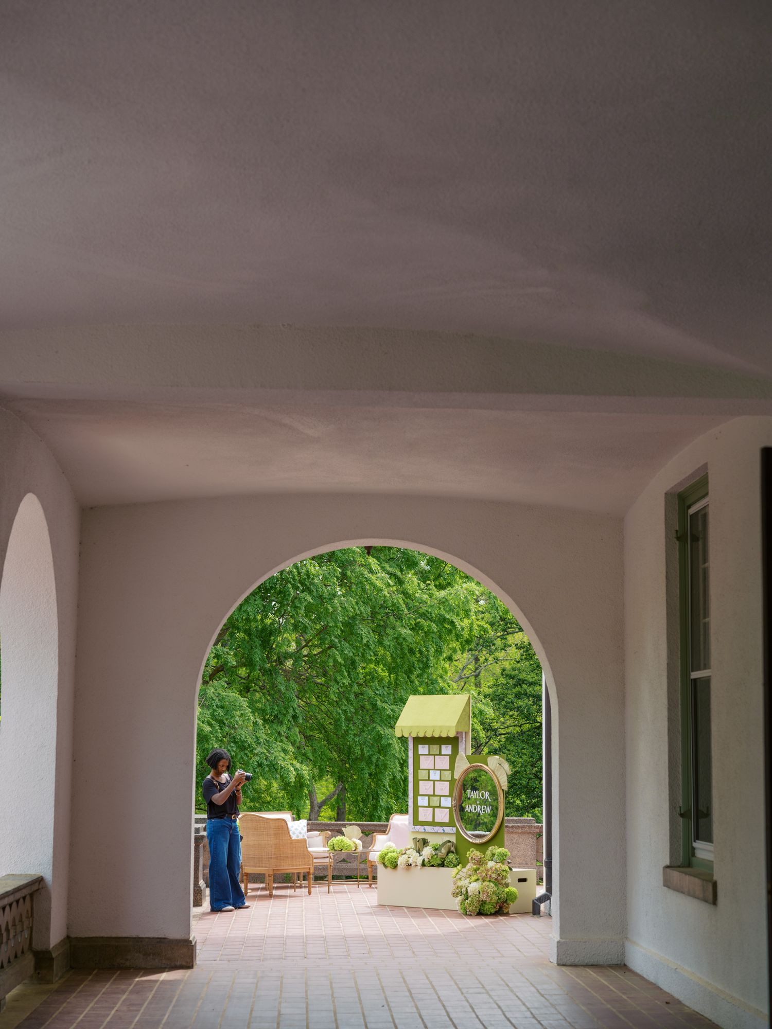 A white arched passageway frames a lush green garden courtyard with a person standing near an informational sign.