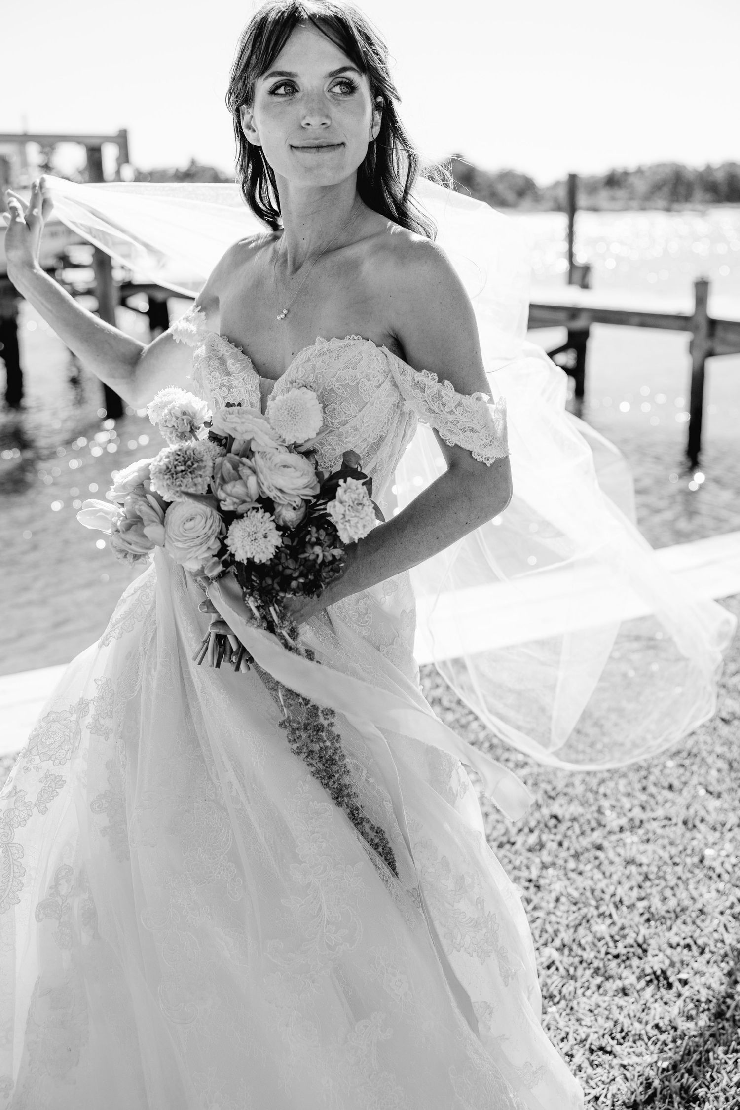 Bride in off-shoulder lace gown holds floral bouquet, veil flowing in wind near waterfront dock.