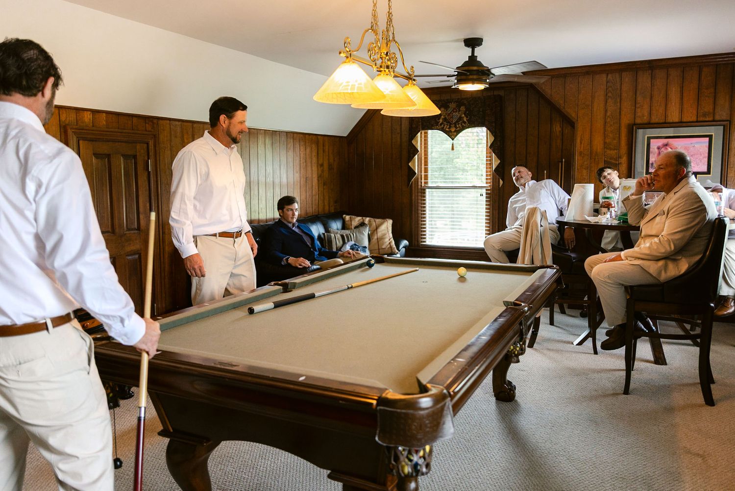 A game room interior with pool tables and wooden paneled walls illuminated by hanging pendant lights.
