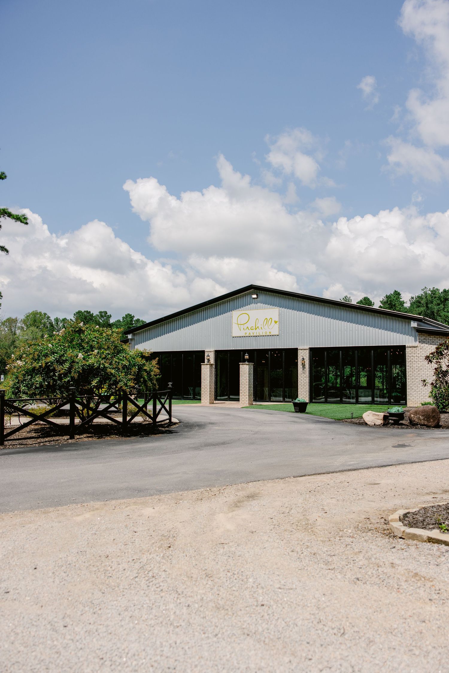 Large commercial warehouse buildings with gravel parking lot and trees under blue sky.