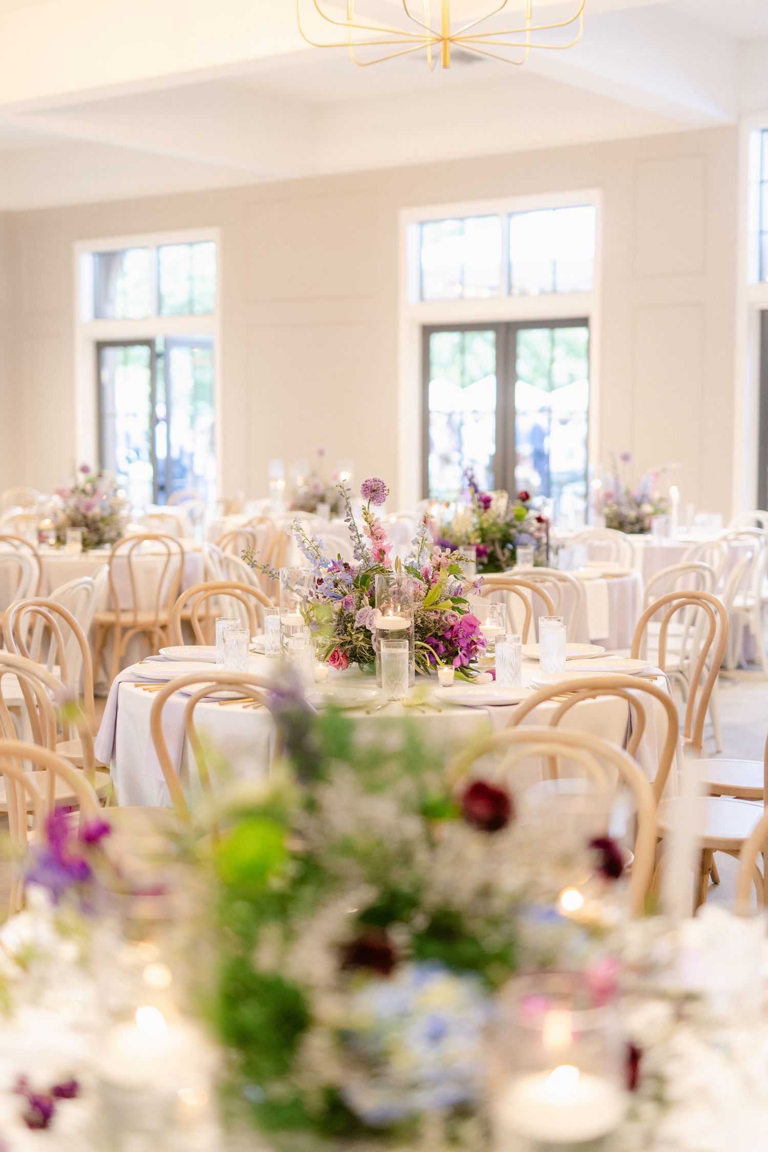 Elegant wedding reception venue with natural light, wooden chairs and delicate floral centerpieces on white tablecloths.