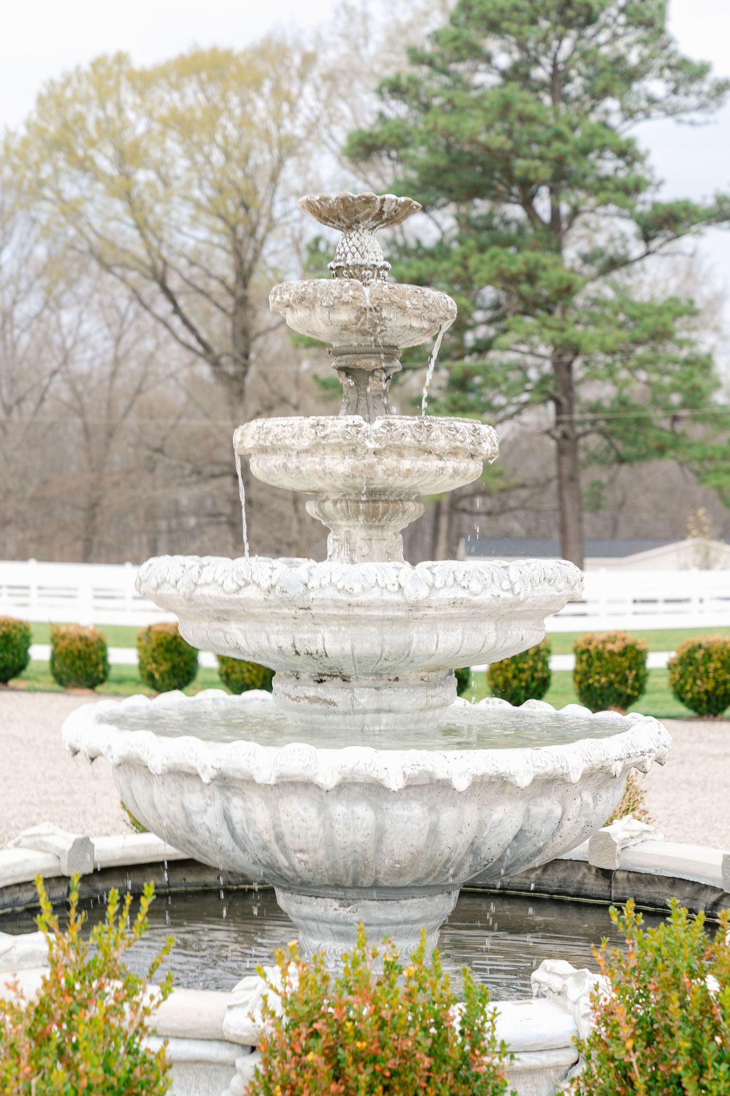 Ornate white stone tiered garden fountain with decorative details surrounded by yellow and orange flowering plants.