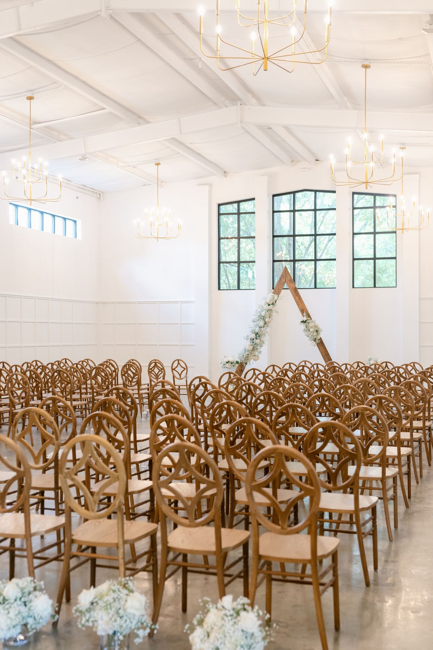 Elegant wedding ceremony setup with rows of wooden chairs in a bright white room with chandeliers and high windows.