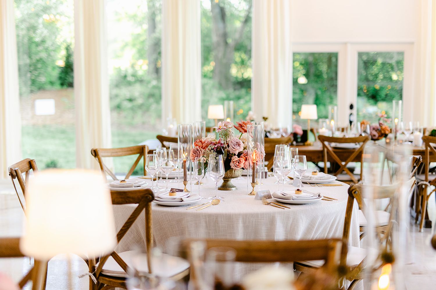 Light-filled reception venue with elegant table settings and cross-back chairs visible through large windows.