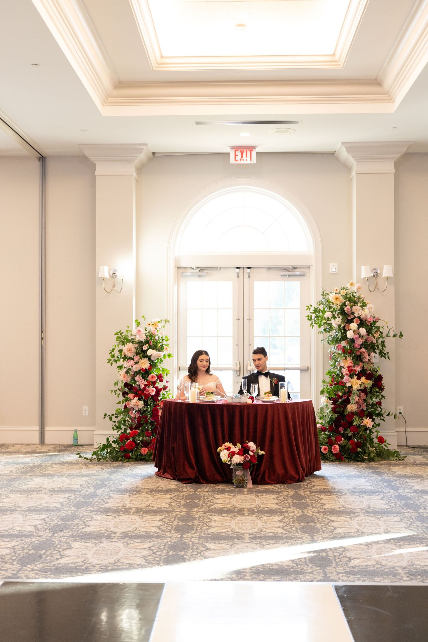 Elegant ballroom setup with burgundy tablecloths and pink floral arrangements along a light gray carpeted floor.