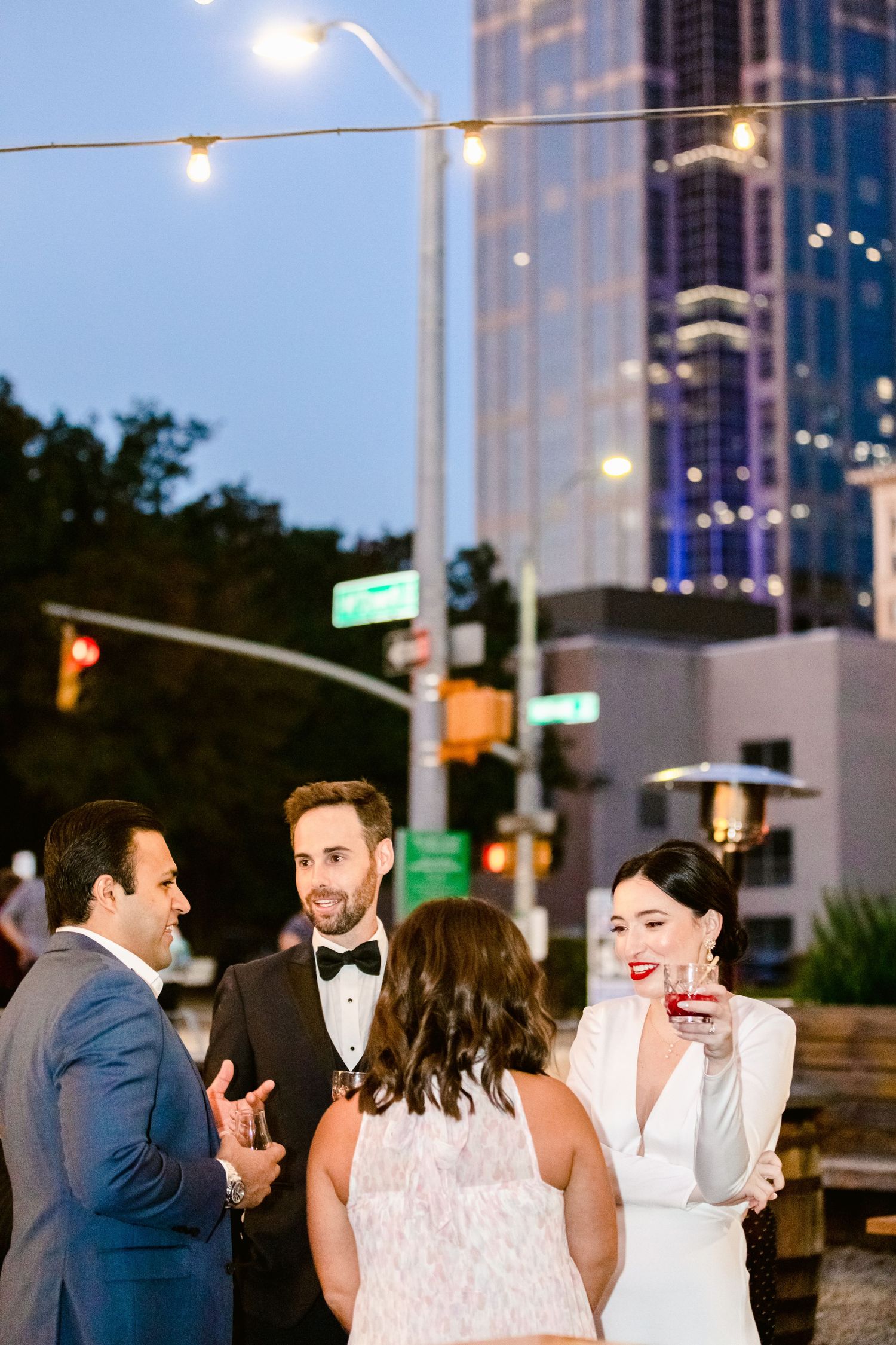 Wedding guests mingle and socialize at an outdoor evening reception with city buildings in the background.