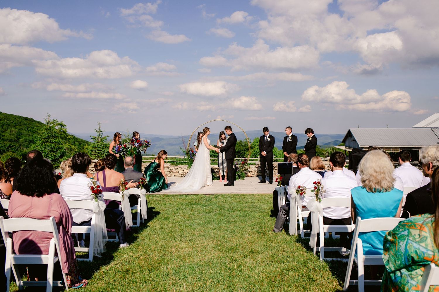Scenic hilltop wedding ceremony with guests seated in white chairs under blue sky.