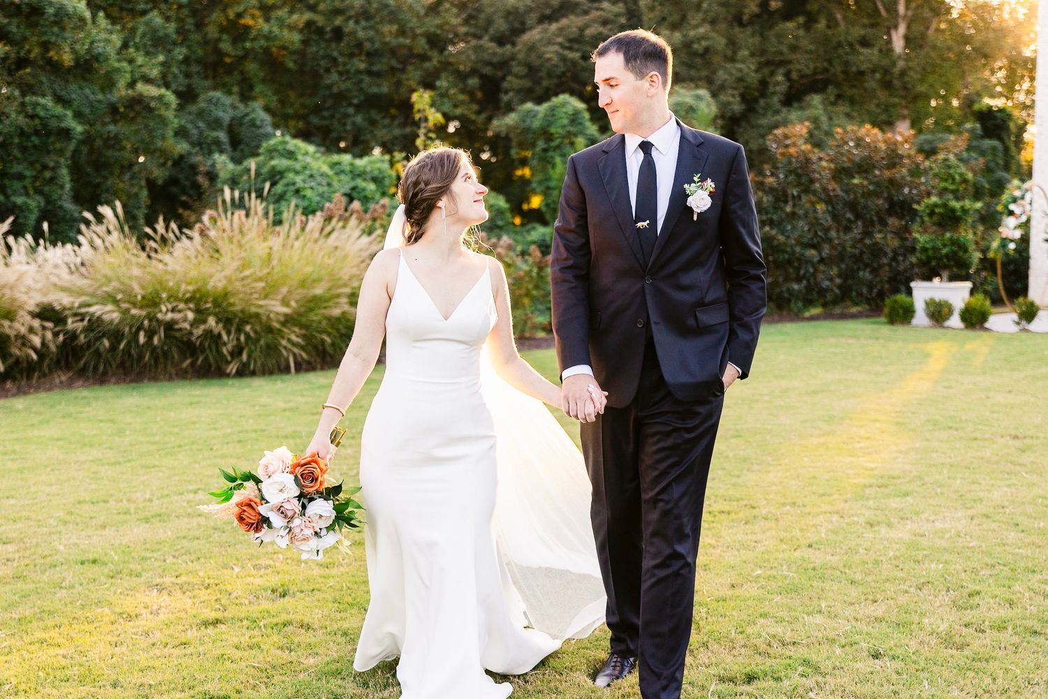 A blissful wedding couple walking hand in hand on a sun-drenched lawn while the bride holds a vibrant bouquet.