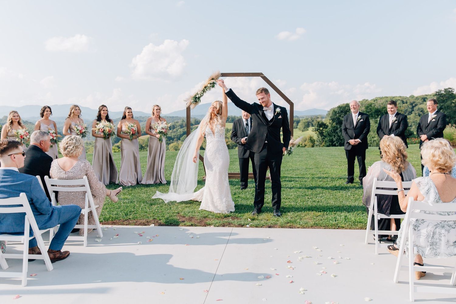 Outdoor wedding ceremony on grassy hilltop with couple under wooden arch and guests seated on white chairs.